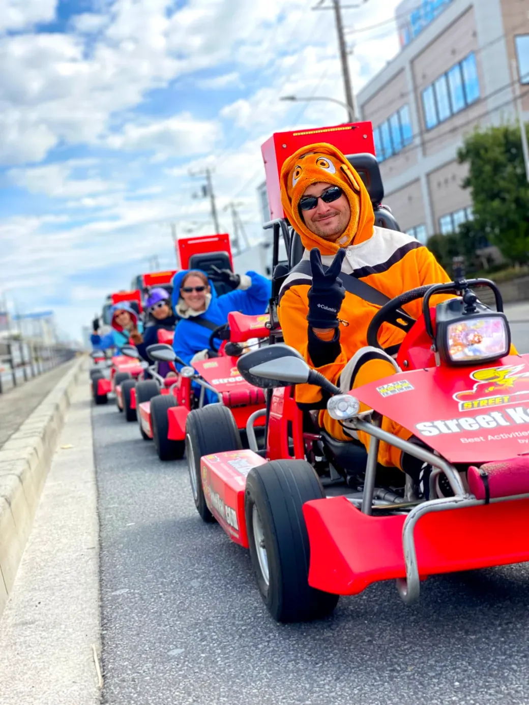 Person in an orange tiger costume on a red go-kart, flashing a peace sign as others ride in a line along a city street.
