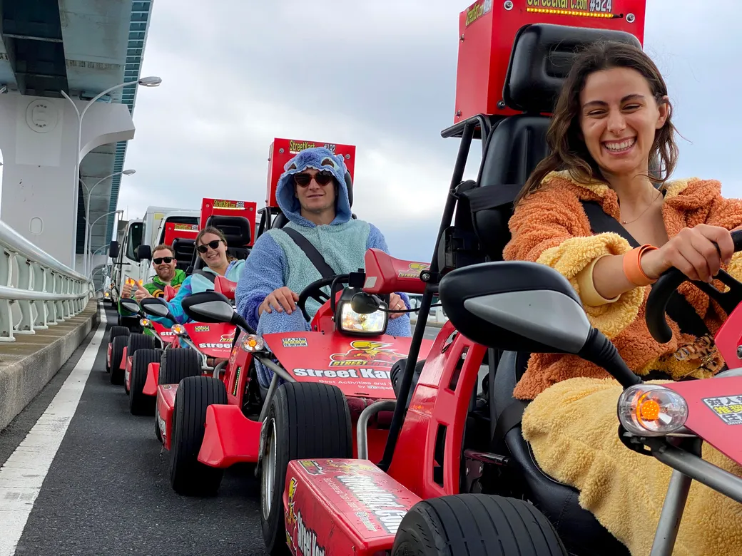 Group of friends in red go-karts lined up on a bridge, smiling and wearing colorful outfits under an overcast sky.